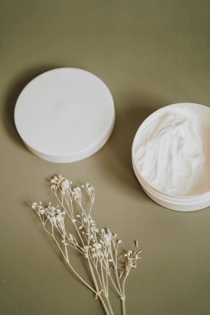 Natural skincare cream in white jar with delicate flowers on green background.